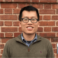 Researcher Quan Zeng standing in front of a brick wall, wearing black-rimmed glasses and smiling at the camera