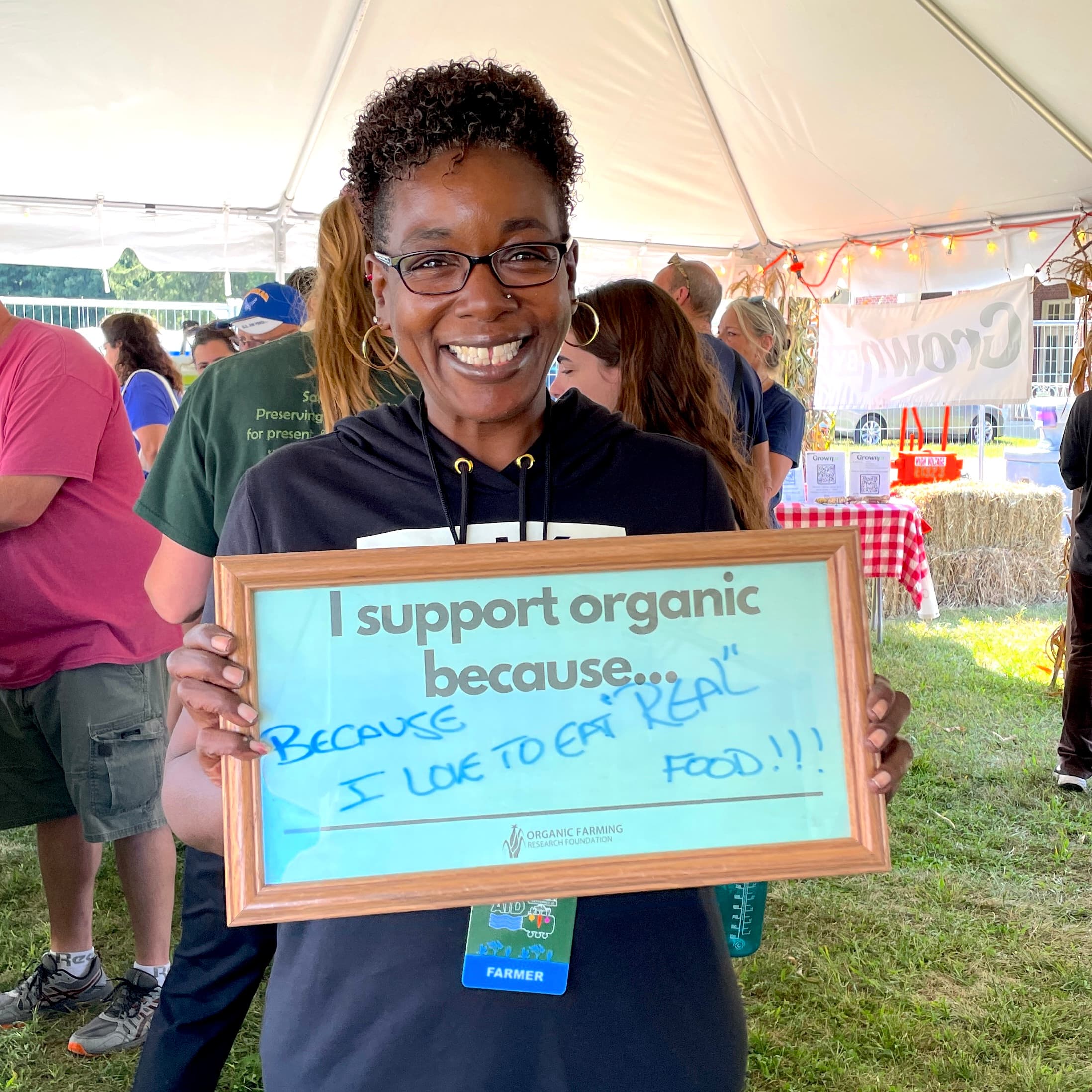 farmer with glasses holding a sign that reads "I support organic because" and she wrote on the blank line "i love to eat 'real' food"
