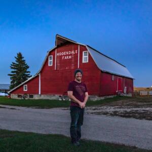 A farmer poses in front of a red barn at dusk.